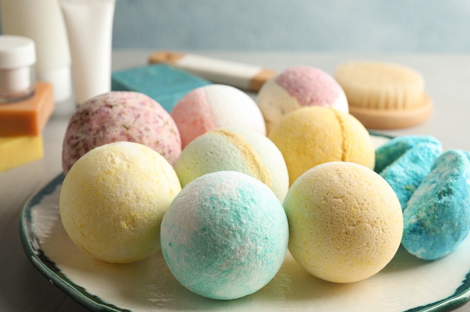 A variety of colorful bath bombs are arranged on a plate. In the background, spa accessories like a brush and soap create a calm, soothing atmosphere.
