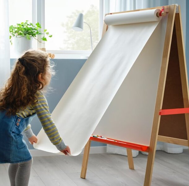 A little girl stands beside a whiteboard, smiling and pointing at drawings on the board.