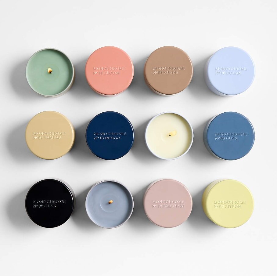 Twelve round candles in pastel tins arranged in a grid. Each tin is a different color, including green, pink, tan, blue, navy, and yellow. Calm and organized.