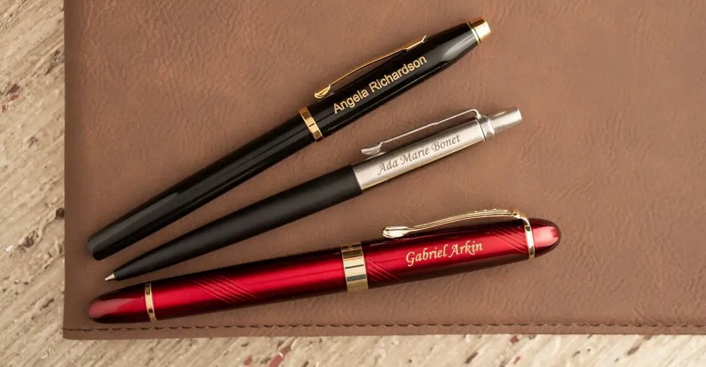 Three pens with engraved names lie on a leather surface: a black pen, a silver pen, and a red pen. The setting conveys elegance and personalization.