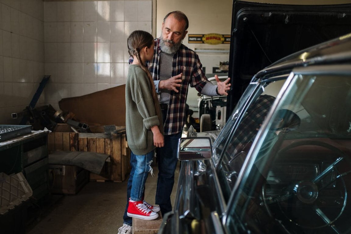A man and his daughter examine a car parked in a garage, both showing interest in the vehicle's features.