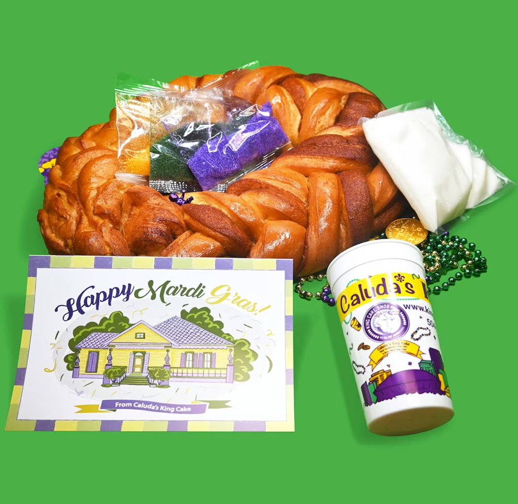 A braided King Cake with icing and sugar packets is surrounded by Mardi Gras beads. A festive "Happy Mardi Gras" card and themed cup complete the scene.