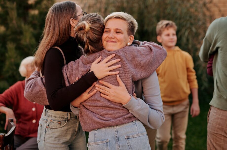 Two women embrace warmly in front of a group of people, conveying a sense of friendship and support.