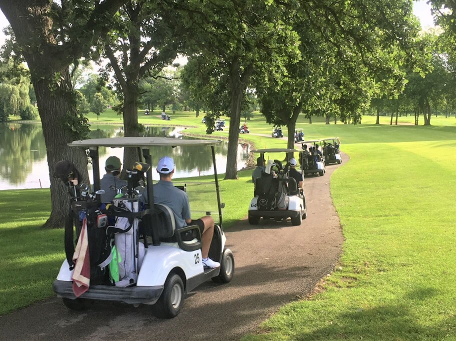 Golf carts with players drive along a tree-lined path beside a tranquil lake on a sunny day, conveying relaxation and outdoor enjoyment.