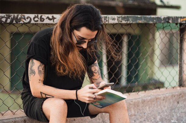 A man with visible tattoos sits on a wall, engaged with his smartphone, appearing relaxed and casual.