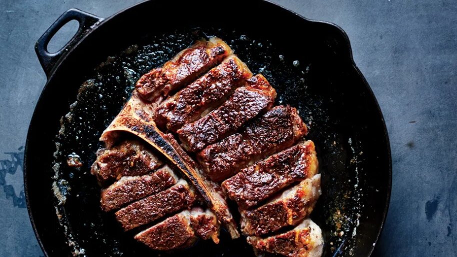 A juicy, seared steak sliced into pieces is shown in a black skillet on a dark surface. The well-cooked meat has a rich, brown texture.