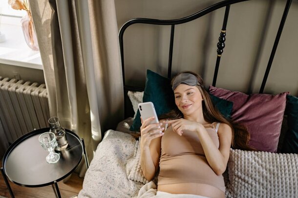 A pregnant woman lying in bed, looking at her phone with a relaxed expression.