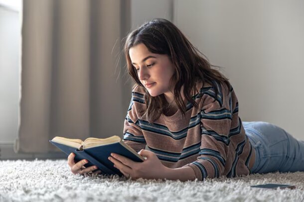 A woman lies on the floor, engrossed in reading a book, surrounded by a cozy atmosphere.