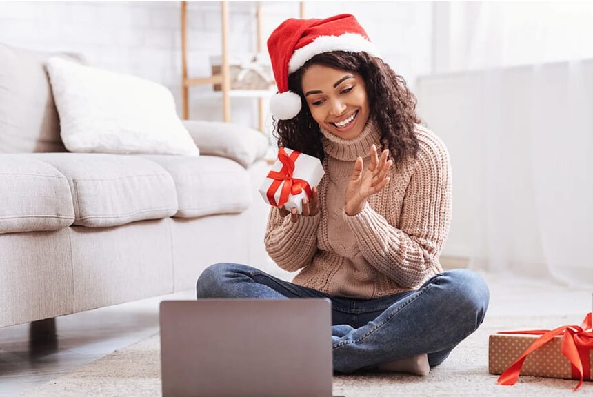 A woman wearing a Christmas hat sits on the floor, focused on her laptop in a cozy holiday setting.