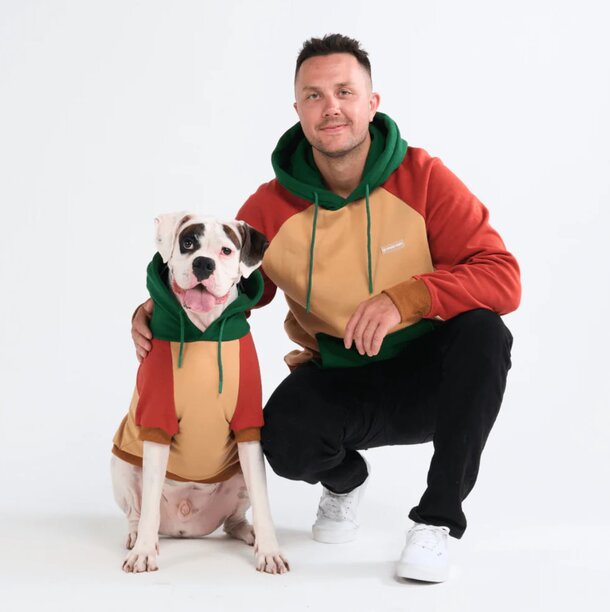A man and his dog, both wearing matching hoodies, stand together in a casual outdoor setting.