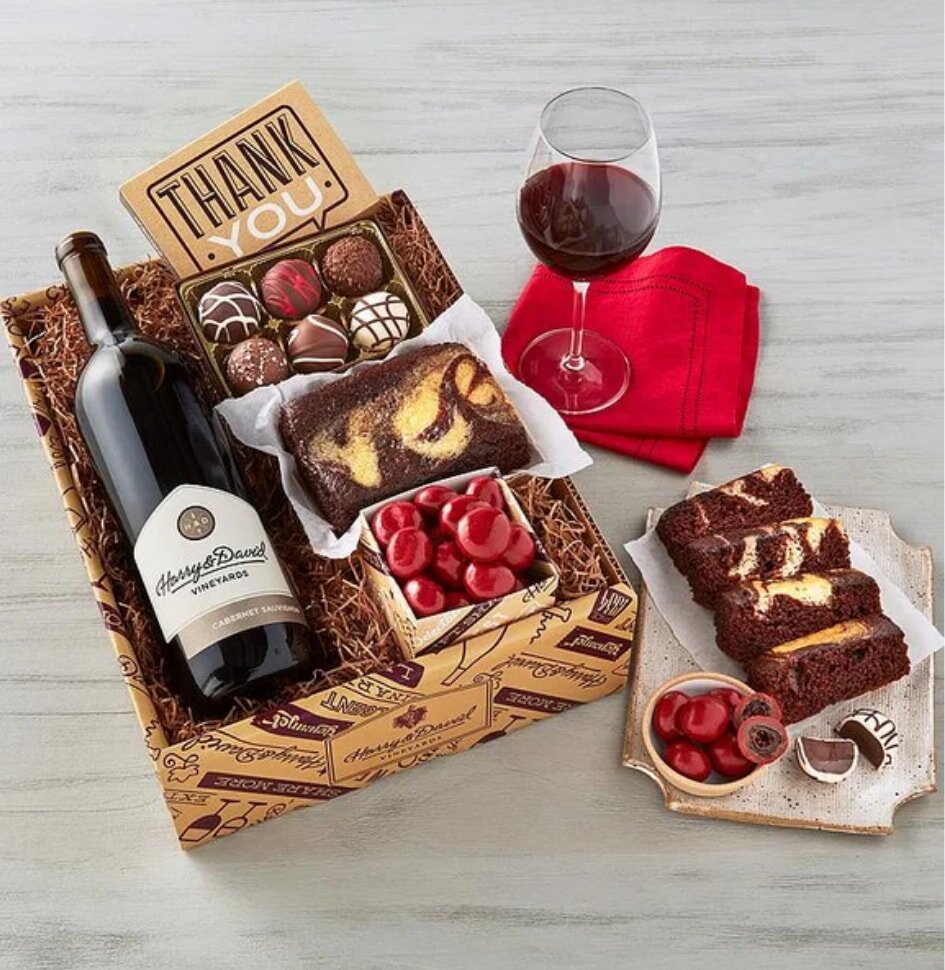 Gift box with a wine bottle, assorted chocolates, a loaf cake, and cherries. A glass of red wine sits beside a red napkin, creating a cozy, inviting scene.