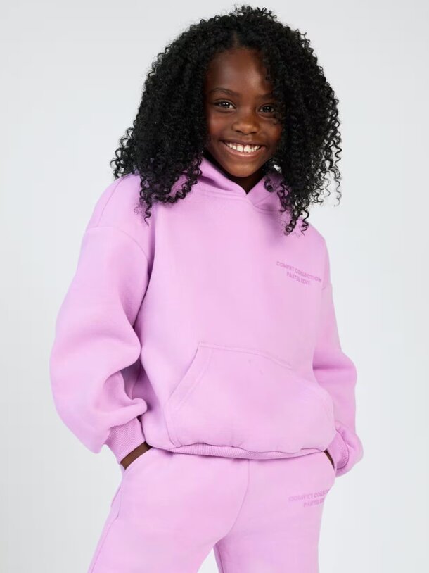 A young girl in a purple sweatshirt and sweatpants stands smiling, showcasing her casual and comfortable outfit.