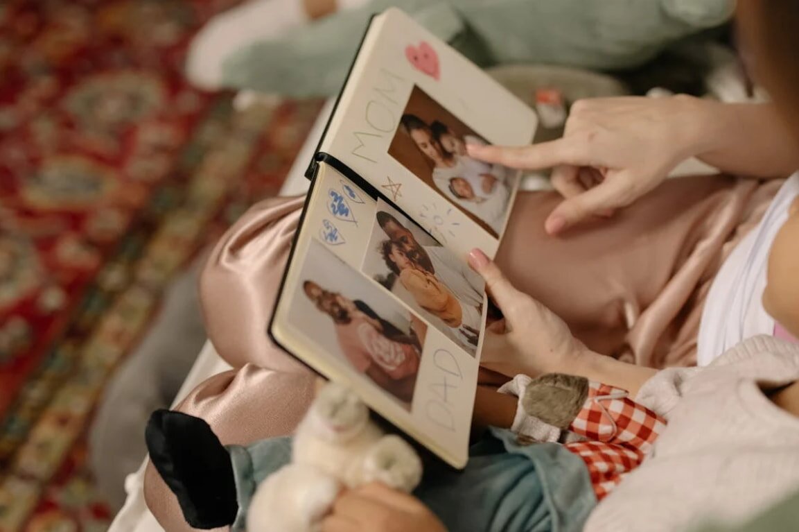 A woman holds a baby in one arm while reading a book with the other hand, smiling gently at the pages.