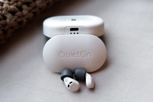 White noise-canceling earbuds with a sleek, oval case on a textured surface. The "QuietOn" logo is visible, conveying calm and technology.