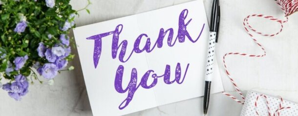 A card with "Thank You" written in purple script lies on a marble surface. Nearby are purple flowers, a pen, and red-striped gift wrap, conveying gratitude.
