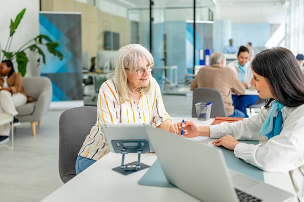 An older woman engages with a female advisor in a modern office. The atmosphere is collaborative, with colleagues in the background, conveying a professional tone.
