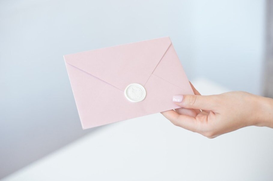 A hand holds a pale pink envelope sealed with a white wax stamp. The soft colors and clean background convey elegance and simplicity.