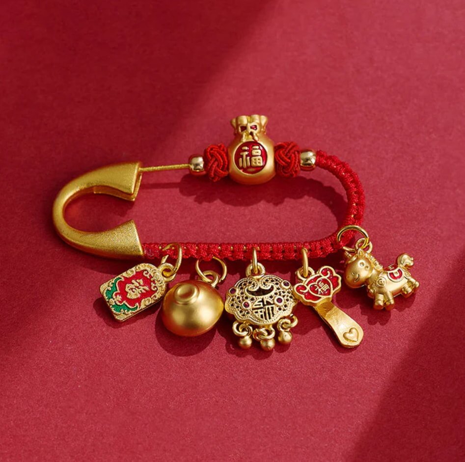 A decorative gold safety pin with red string holding six gold charms, including a bag, coins, and a horse, on a rich red background, conveying luck and prosperity.