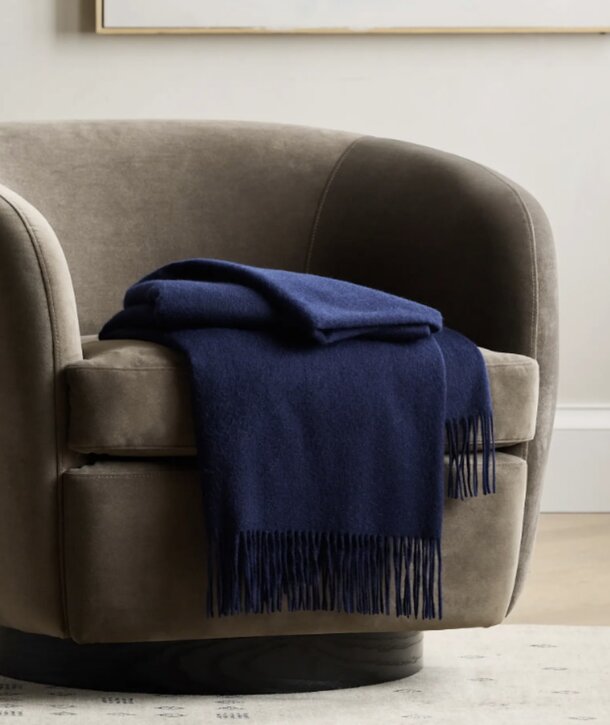A plush, taupe velvet armchair is draped with a soft, fringed navy blue throw blanket, creating a cozy and elegant atmosphere in a minimalist room.