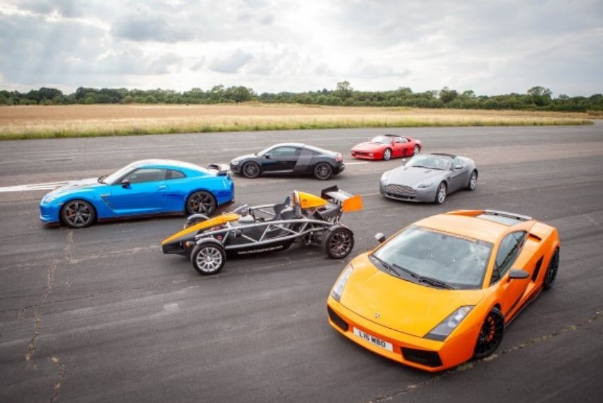 Five sports cars, including an orange and blue one, are parked on an open runway. The scene is set on a cloudy day, conveying a dynamic feel.
