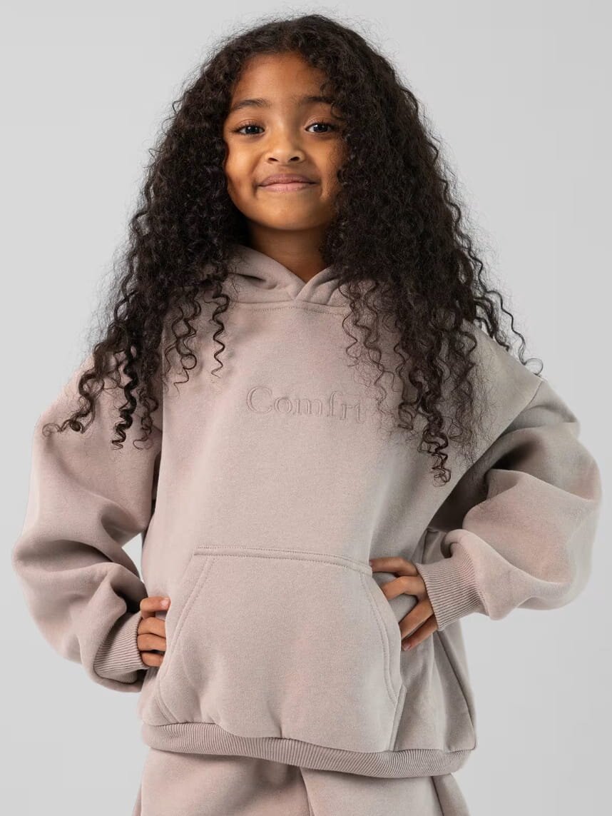 A young girl in a grey hoodie and sweatpants stands, smiling, with her hands in her pockets.