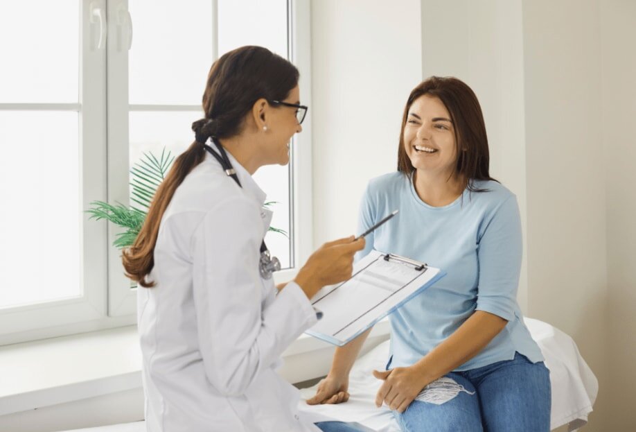 A doctor in a white coat and glasses consults with a smiling patient in a light blue shirt. They sit near a window, creating a warm, friendly atmosphere.