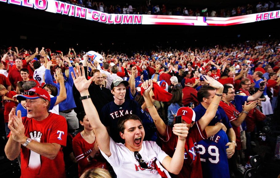 A cheering crowd at a baseball game, wearing predominantly red and blue team apparel. The atmosphere is energetic and celebratory, with people clapping and raising hands.