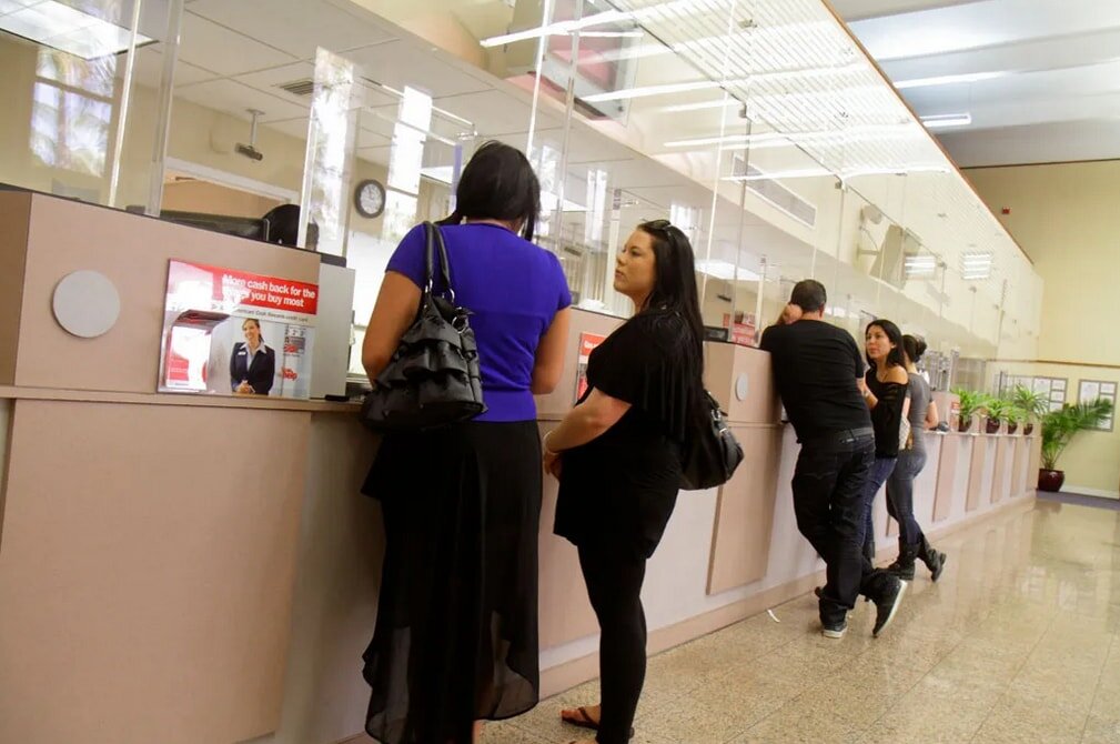People stand in line at a bank counter with glass partitions. The atmosphere is busy yet calm, with potted plants and bright lighting.