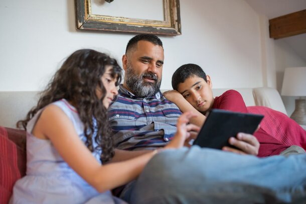 A man and two children are sitting on a couch, engaging with a tablet together.