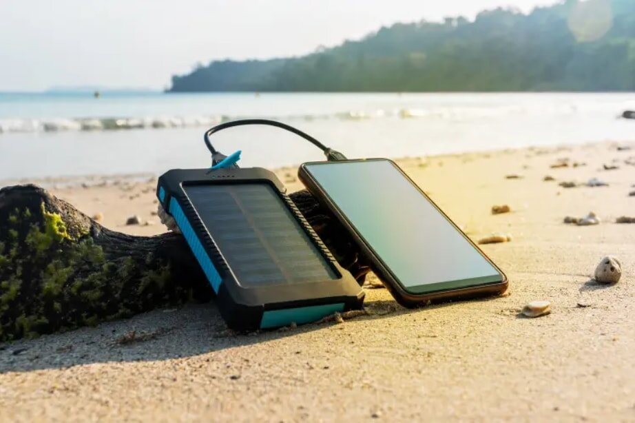 A smartphone and solar charger lie on sandy beach near driftwood, with waves and sunny, distant hills in background. Tech and nature in harmony.