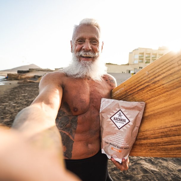 Older fit man taking a selfie on the beach with his surfboard and bag of Ka'Chava superblend.