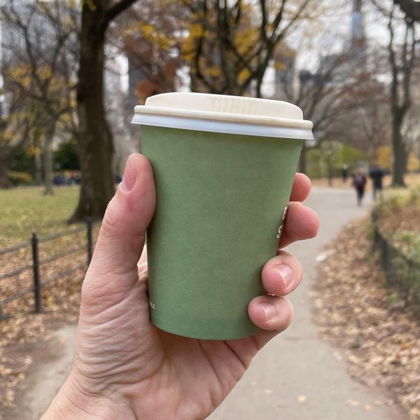 Green paper coffee cup held aloft before a park scene.