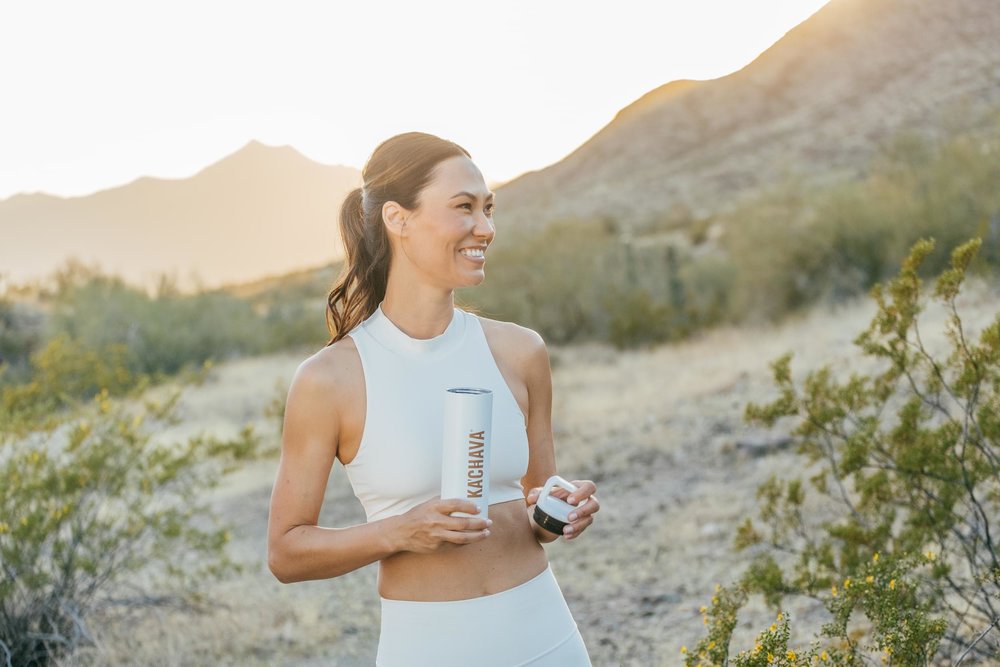 Fit young woman in the outdoors at sunrise, smiling to her right and drinking Ka'Chava.