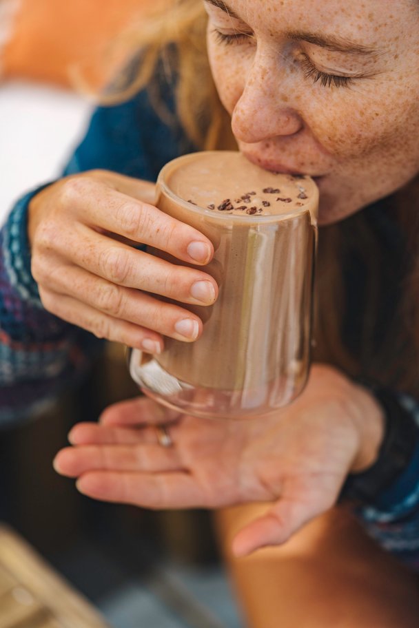Freckled woman takes a sip of Ka'Chava chocolate shake from an over-flowing cup.