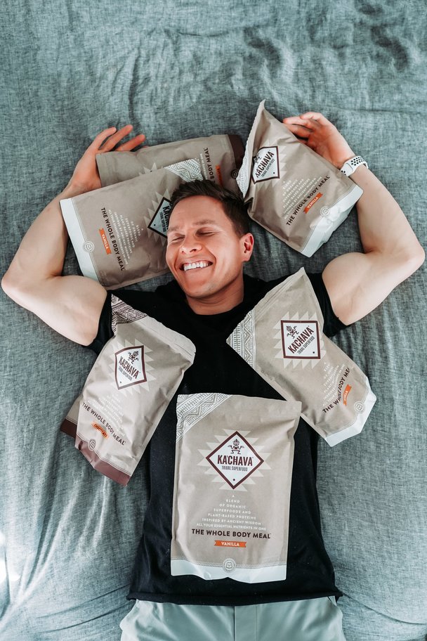 Fit man laying on a soft surface, smiling and surrounded by bags of Ka'Chava superblend.