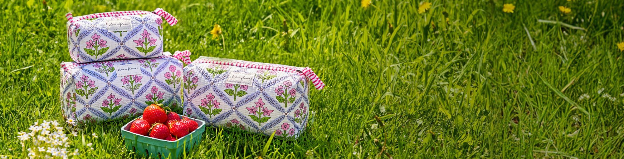Strawberry-print quilted Strawflower set of 3 cosmetic pouches and fresh strawberries arranged in tall green grass on a bright summer day.