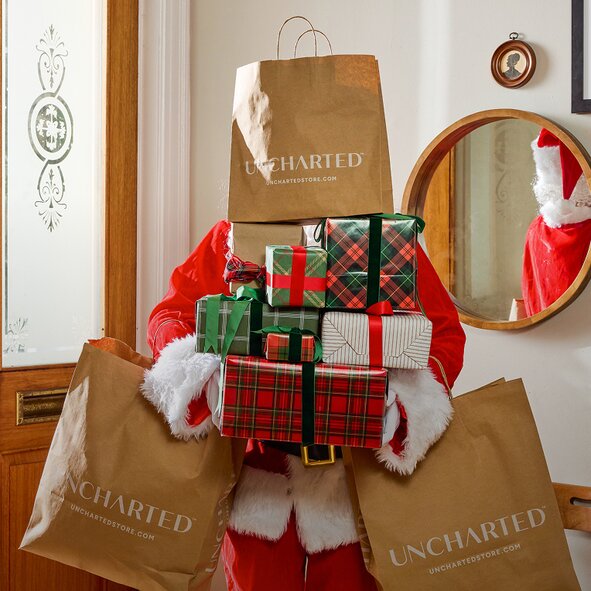 Santa carrying a stack of wrapped gifts and Uncharted shopping bags inside a home.