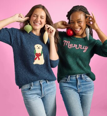Two girls wearing holiday sweaters holding ornaments up to their ears as if they were earrings
