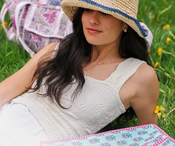 A woman relaxes in a flower-filled grassy field beside colorful quilted tote bags and a matching open tote, wearing a straw sun hat and white Free People tank.