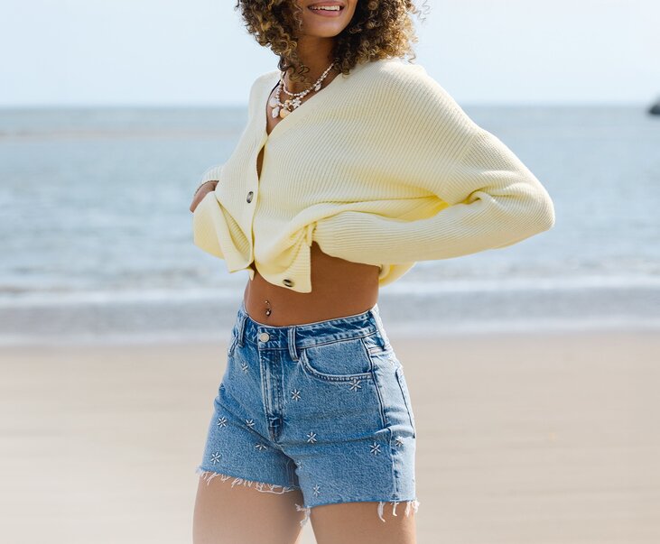 A woman stands on the beach wearing a pale yellow knit cardigan, embroidered denim shorts, and layered shell necklaces, with the ocean in the background.