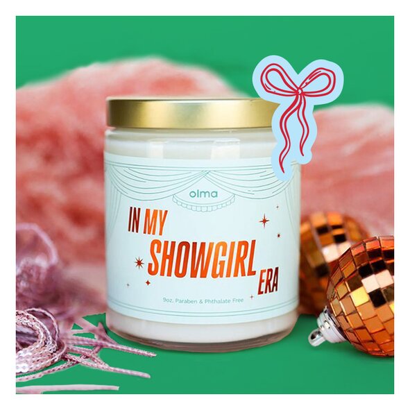 Glass candle labeled "IN MY SHOWGIRL ERA" with a gold lid and light blue label 