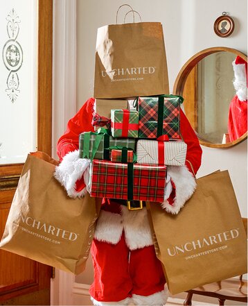 Santa carrying a stack of wrapped gifts and Uncharted shopping bags inside a home.
