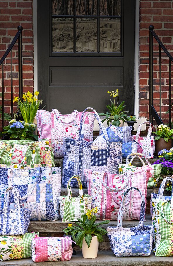A collection of colorful, quilted patchwork Strawflower tote bags and small pouches arranged on stone steps in front of a black door and red brick house, surrounded by blooming spring flowers in pots.