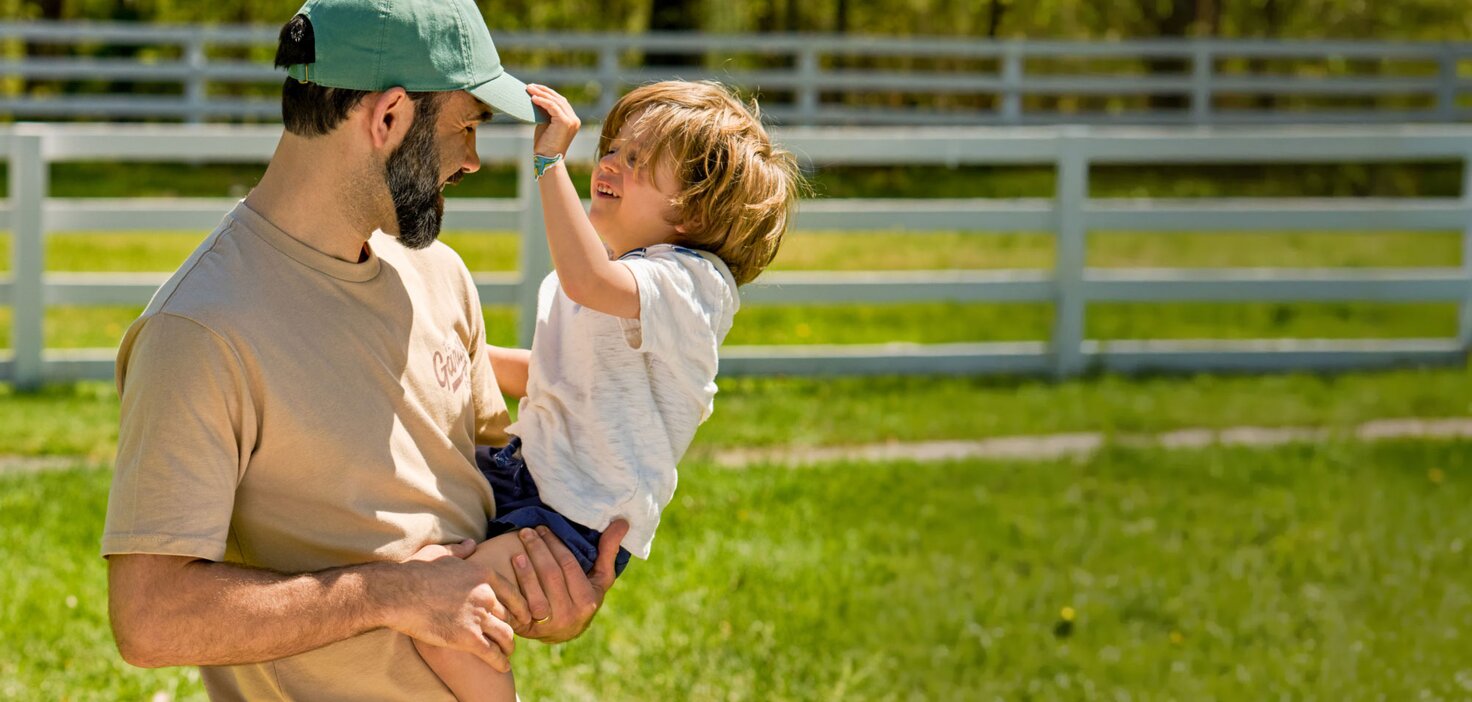 Father and child outdoors in a sunny grassy field, with the child reaching up to touch the man’s green baseball cap near a white fence.