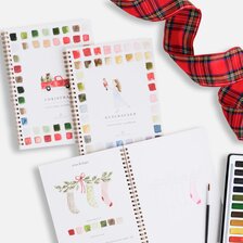 Christmas-themed Emily Lex watercolor workbooks featuring festive illustrations and color palettes, arranged with watercolor paints, a brush, and red plaid ribbon.