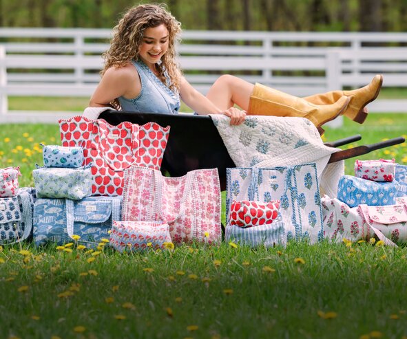 Woman sitting on grass behind a collection of floral and patterned Strawflower tote bags, duffels and cosmetic bags.