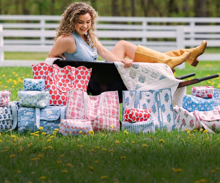 Woman sitting on grass behind a collection of floral and patterned Strawflower tote bags, duffels and cosmetic bags.