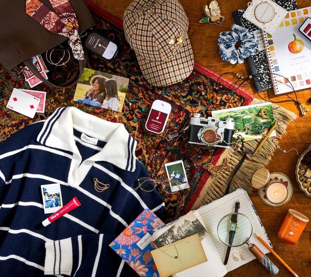 Flat lay of fall accessories and keepsakes, including a striped sweater, plaid hat, jewelry, photos, notebooks, candles, and Touchland hand sanitizers arranged on a patterned rug and wooden floor.