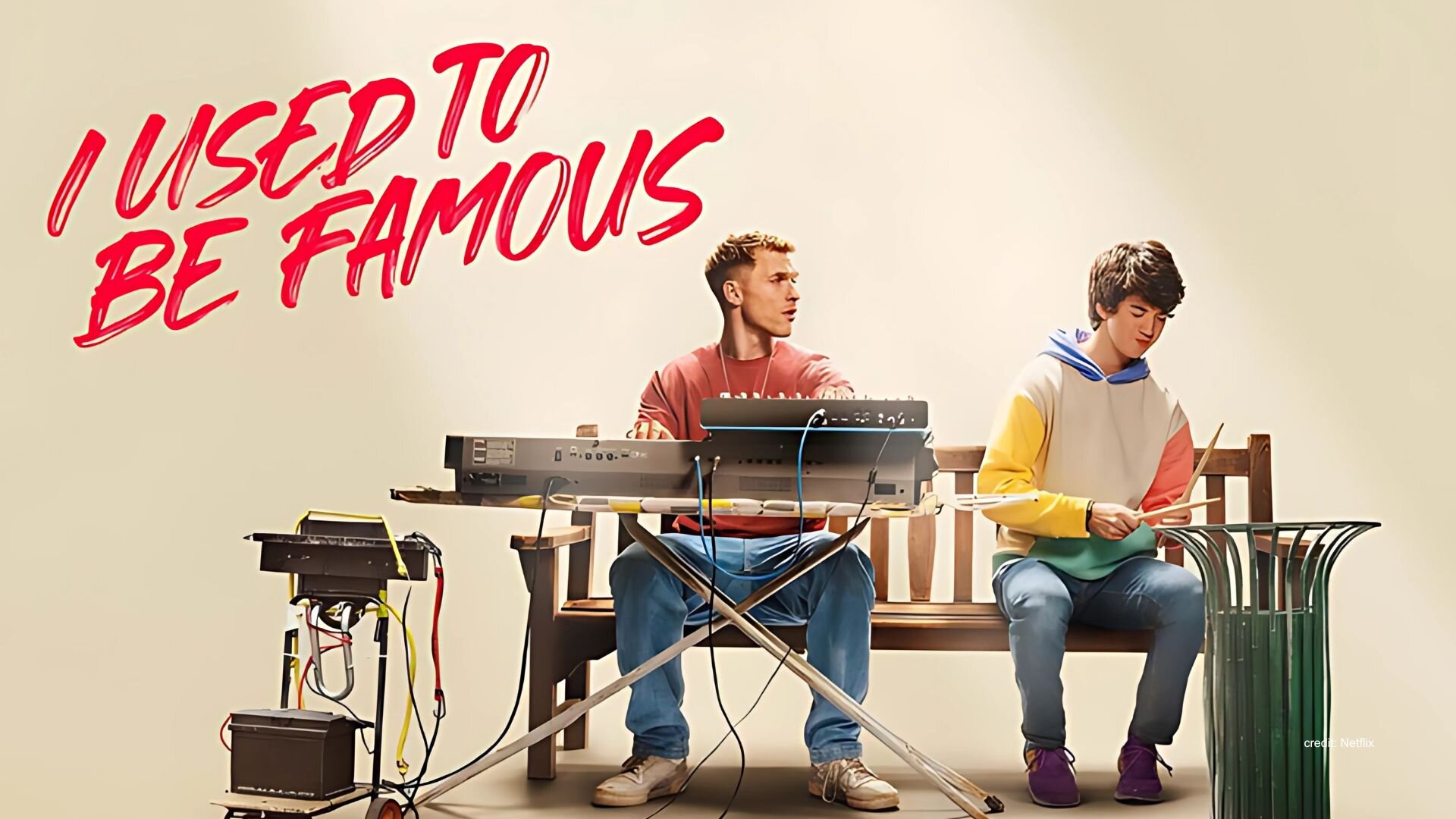 Promotional image for the Netflix movie called I Used To Be Famous. A musician sits on a park bench playing the keyboard in front of him white a young man with autism plays percussion on a green metal garbage can. Red text in the upper left corner reads I Used To Be Famous.