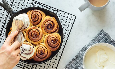 Freshly baked cinnamon buns are being iced in a cast iron pan. 